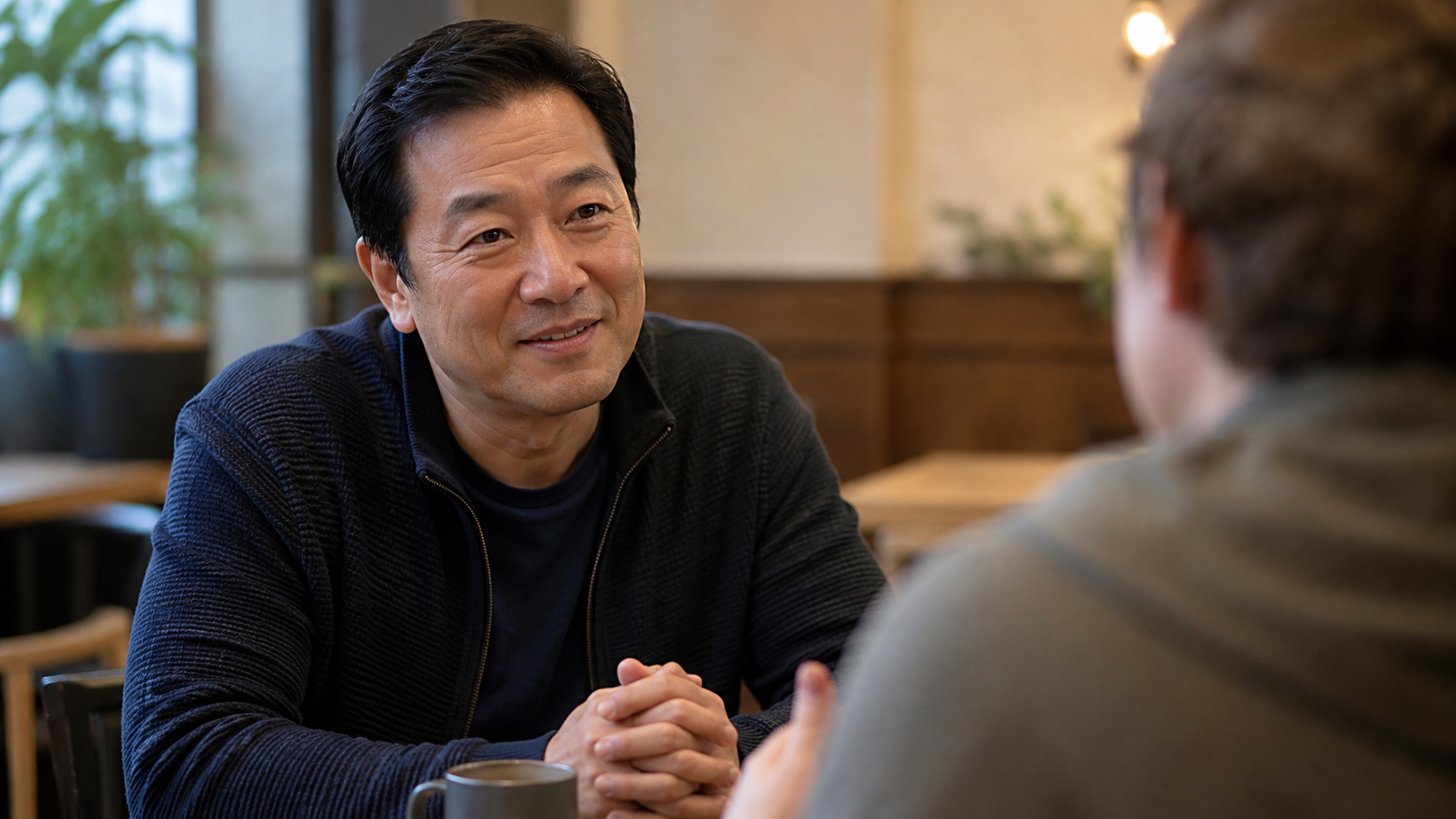 A middle-aged man sitting at a small table leaning in attentively toward another person, representing the power of being truly heard by someone who cares.